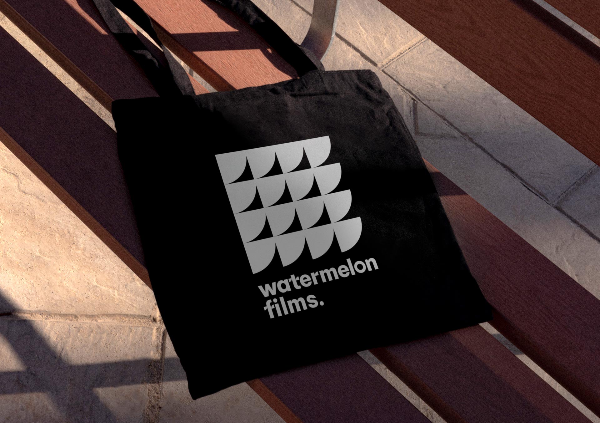 Sac en toile noir avec un design graphique blanc représentant le logo de Watermelon Films, composé d’un motif géométrique et du nom de l’entreprise. Il repose sur un banc en bois avec des ombres créées par la lumière naturelle.